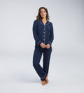 A woman modeling a navy blue two-piece pyjama set with white piping details, consisting of a long-sleeve button-up top and matching pants, against a light grey studio background.