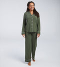 A woman modeling an olive green pyjama set with white piping details, consisting of a long-sleeve button-up shirt and matching wide-leg pants, against a light grey studio background.