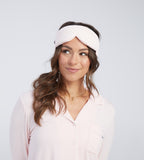 A woman wearing a light pink sleep mask, photographed against a clean light grey studio background.