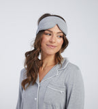 A woman wearing a light grey sleep mask photographed against a clean light grey studio background.
