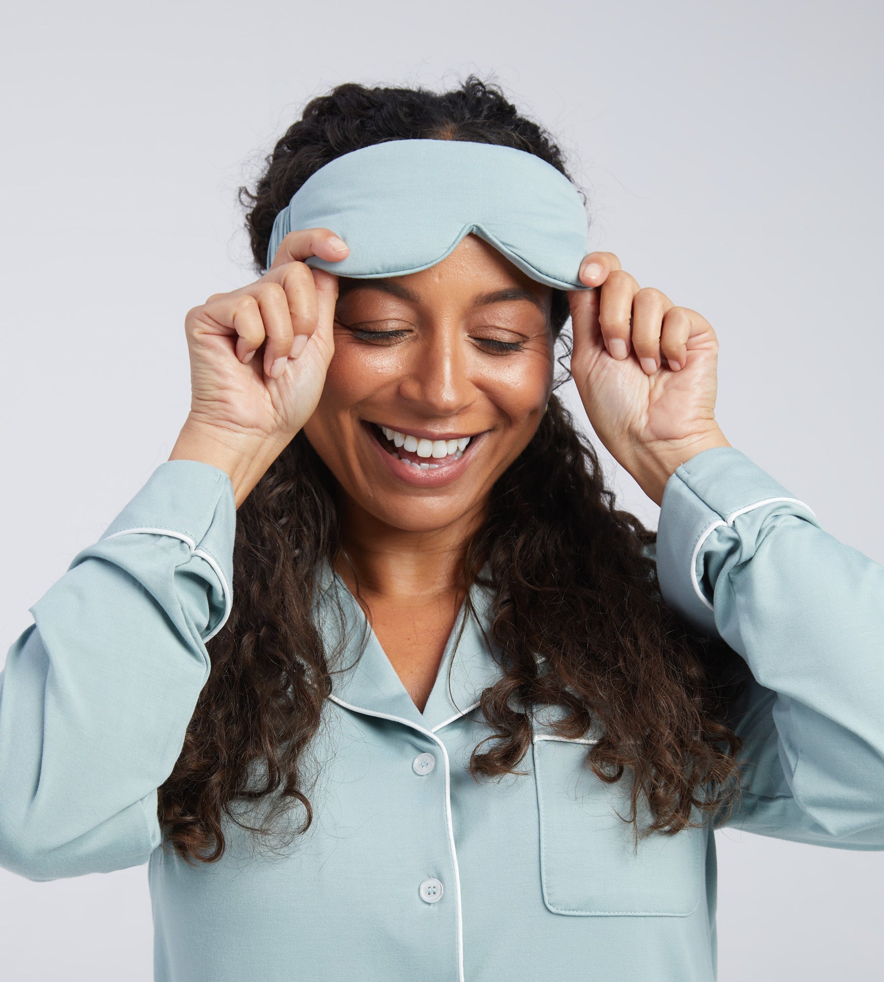 A woman with long curly dark hair wearing a light blue sleep mask, smiling as she adjusts the mask with both hands against a light grey background.