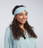 A woman with long curly brown hair wearing a light blue sleep mask photographed against a plain light grey studio background.