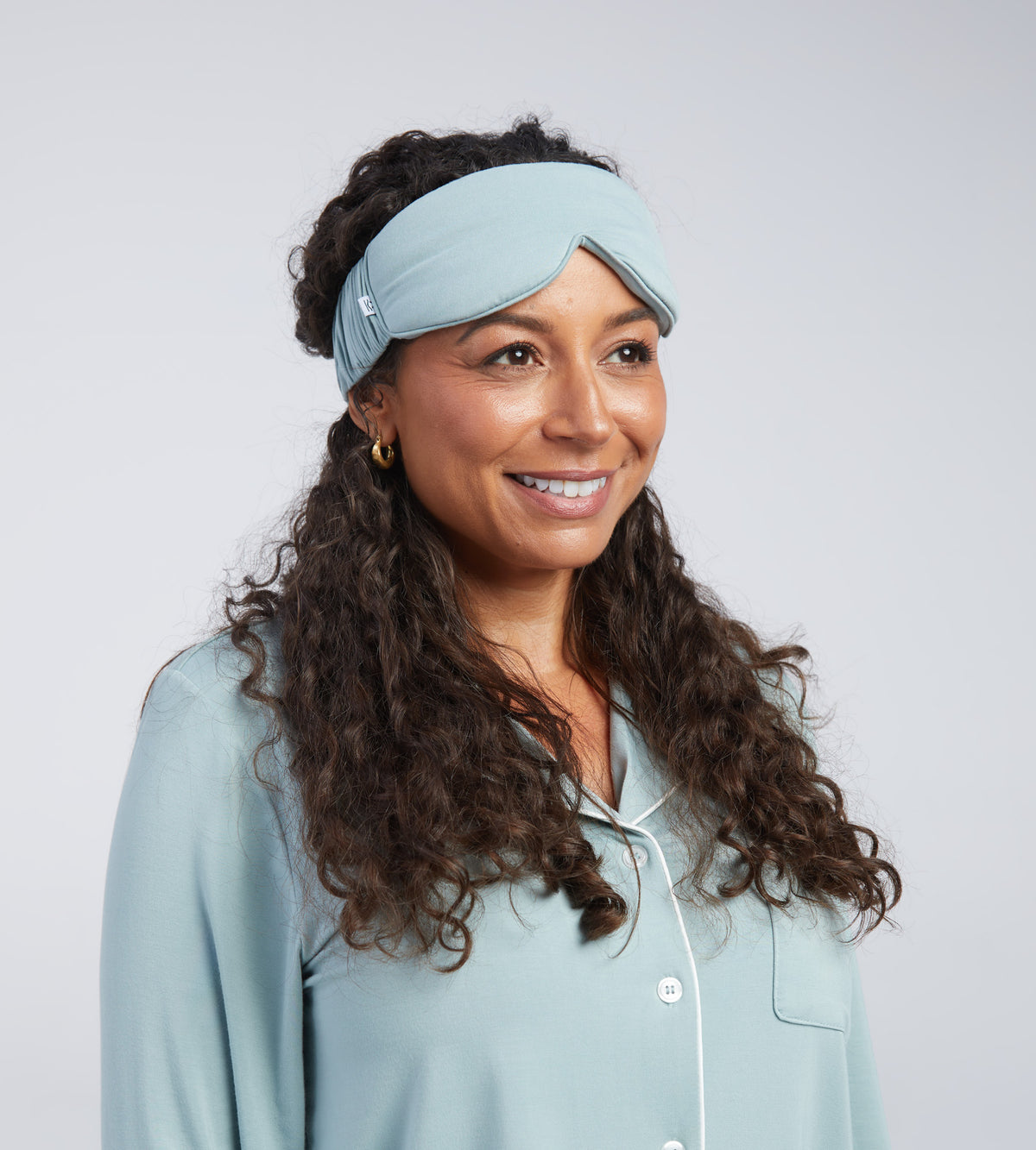 A woman with long curly brown hair wearing a light blue sleep mask photographed against a plain light grey studio background.