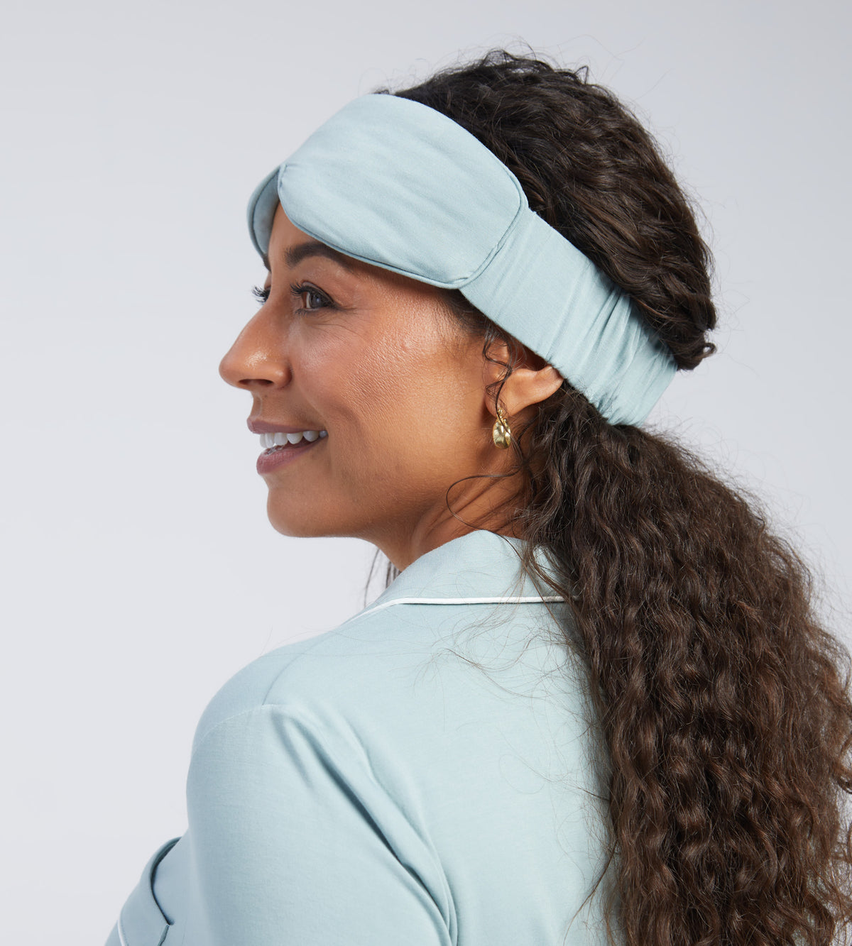 A woman shown in profile wearing a light blue sleep mask against a plain light grey studio background.