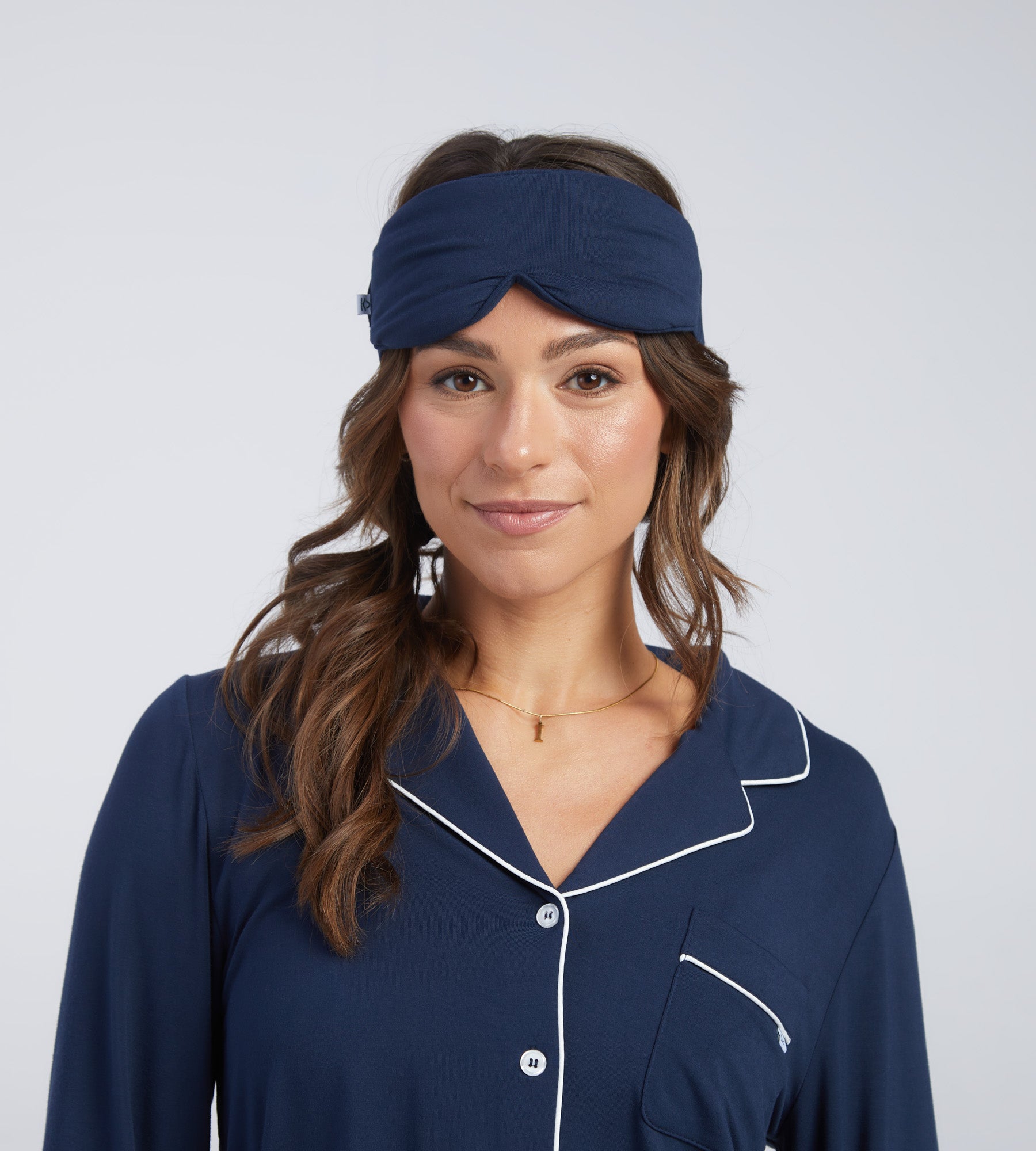 A woman wearing a navy blue sleep mask photographed against a light grey studio background.