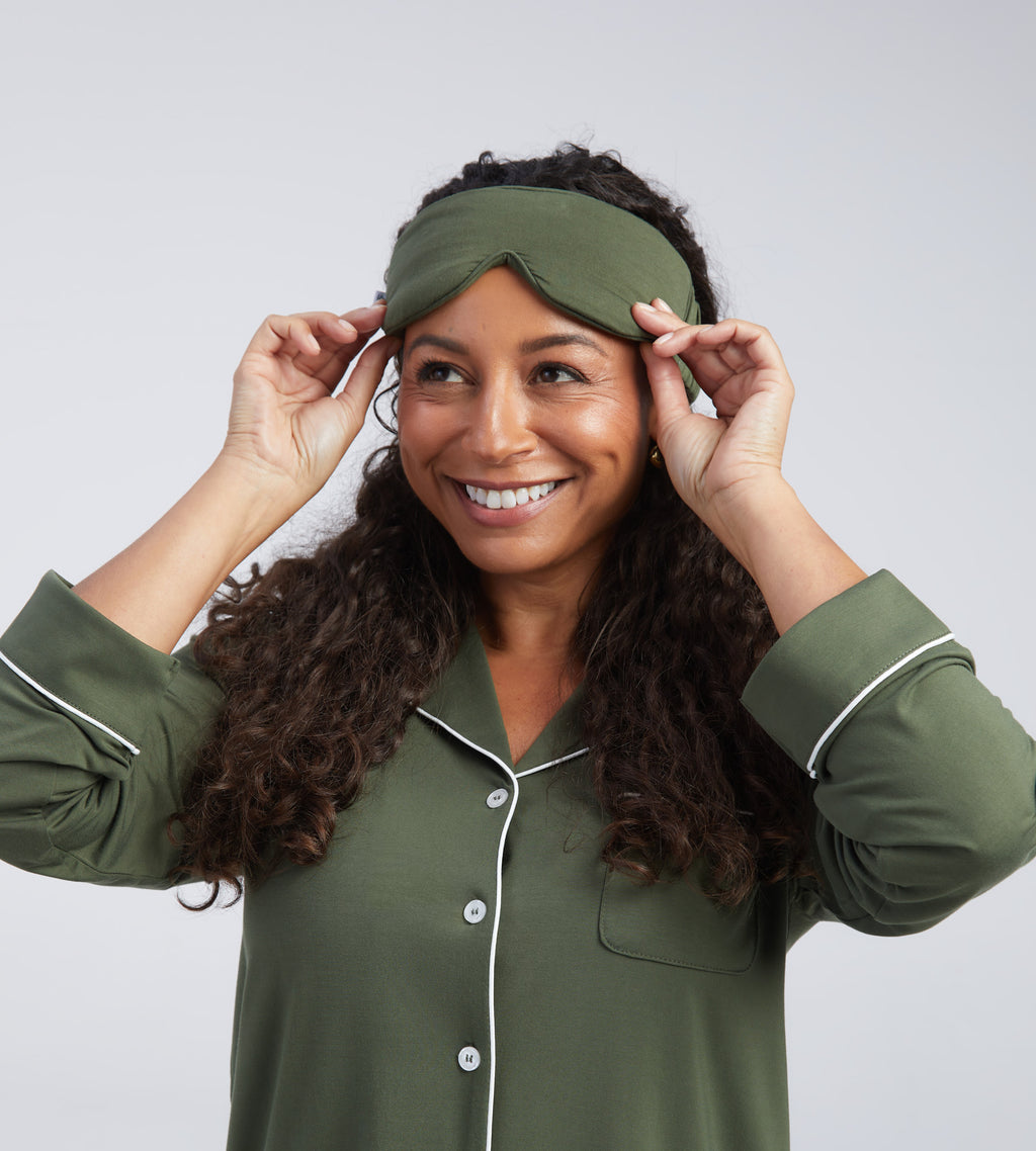 A smiling woman with long curly brown hair wearing an olive green sleep mask adjusting the mask against a light grey studio background.