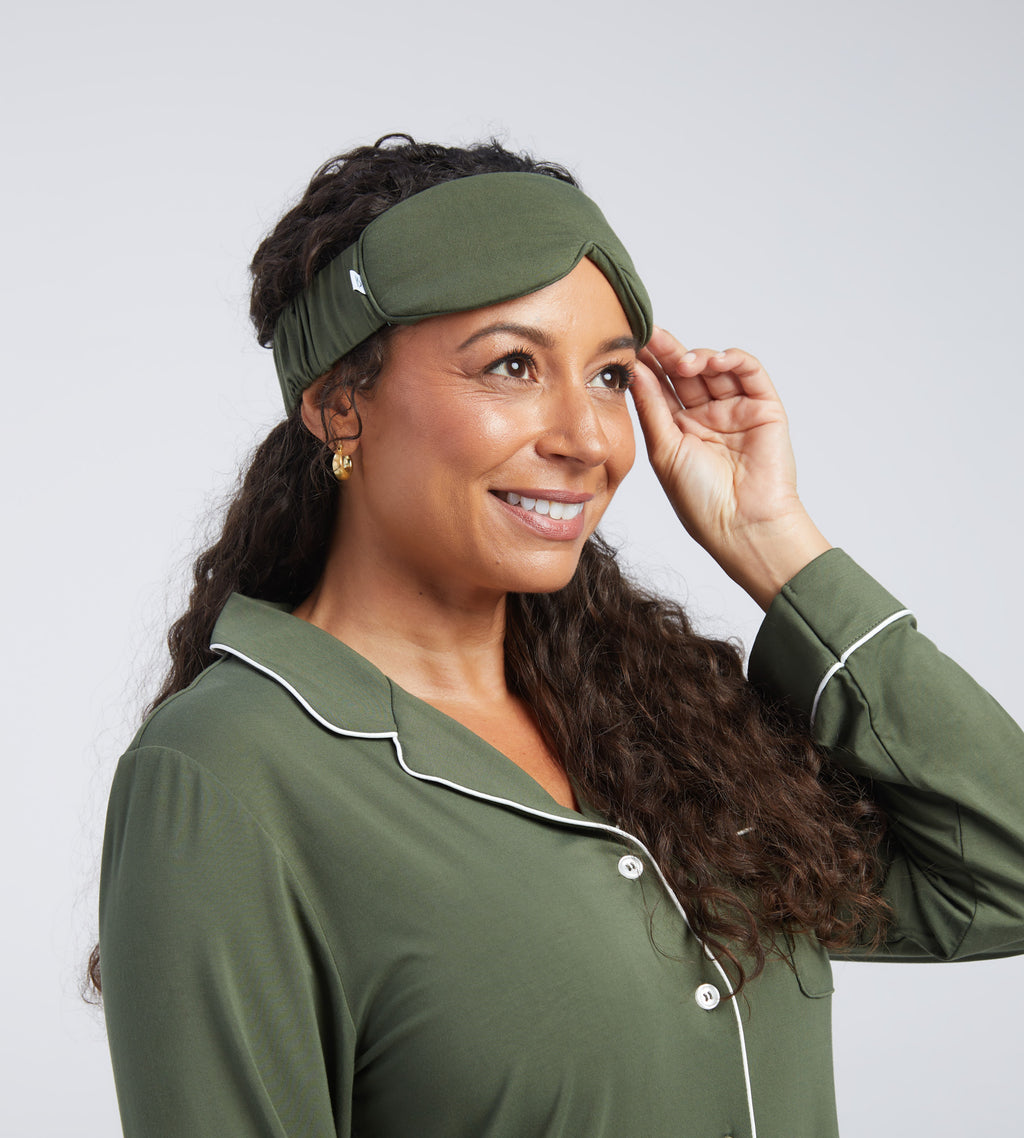 A woman wearing an olive green sleep mask against a light grey studio background.
