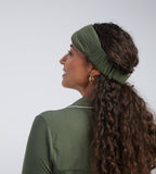 A woman shown from the side and back wearing an olive green wide sleep mask against a light grey studio background.