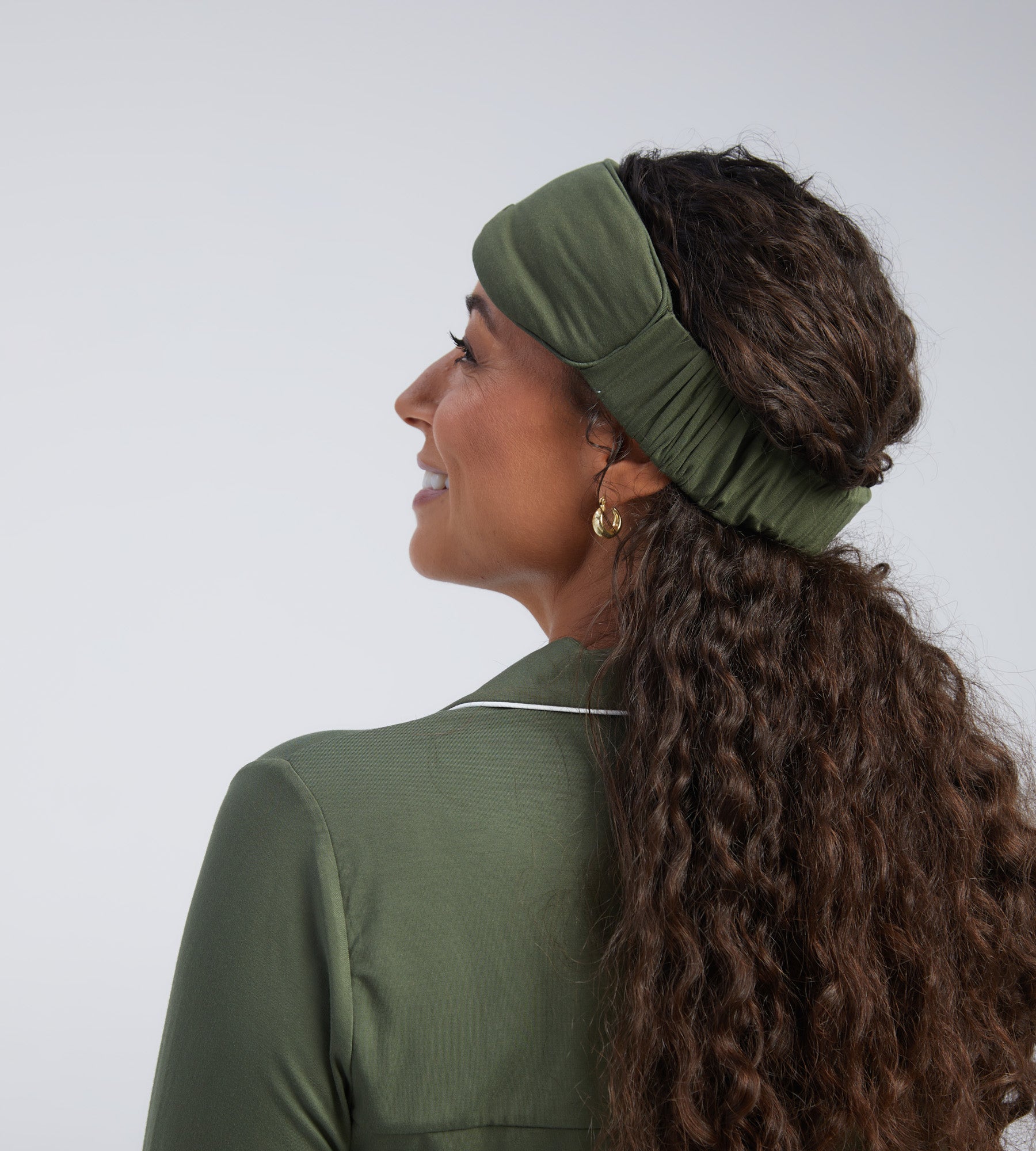 A woman shown from the side and back wearing an olive green wide sleep mask against a light grey studio background.