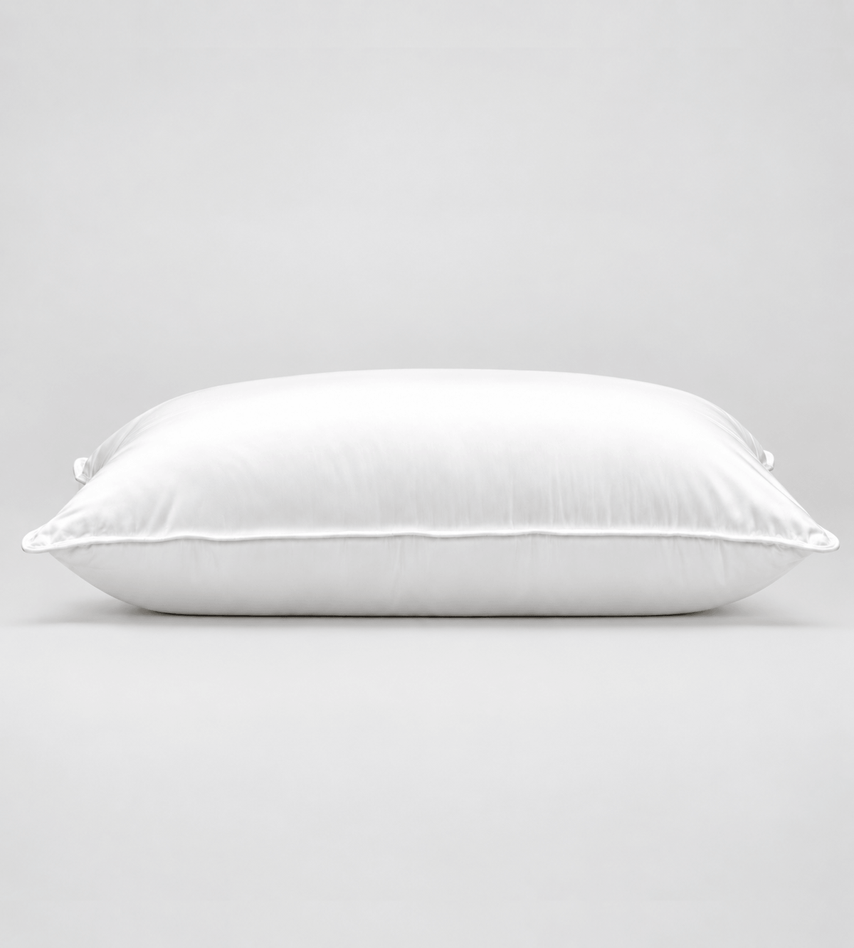 A white rectangular pillow with a smooth, clean surface and piped edges, photographed against a seamless light grey studio background.